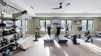 Located on TW Alexander Drive in Durham, NC, this fitness center offers a thoughtfully designed single-floor exercise space with everything needed for a comprehensive workout. The main exercise room is bright and welcoming, filled with abundant natural light streaming through multiple black-framed windows that frame views of an outdoor pool area. Soft sage-green walls, white trim, and a neutral-toned flooring create a clean, modern aesthetic throughout.

The cardio zone features a lineup of treadmills, stationary bikes, and an elliptical machine, each equipped with individual digital screens for interactive workouts. Two wall-mounted flat-screen televisions are positioned for easy viewing from the cardio equipment. Overhead, ceiling fans with sleek, dark-bladed designs provide airflow and comfort during intense sessions.

The strength training area includes a multi-station cable machine and a well-stocked dumbbell rack, complemented by large floor-to-ceiling mirrors accented with bold geometric-patterned wallcovering. The mirrors amplify the sense of space and allow users to monitor their form. Recessed lighting supplements the natural light, ensuring the room remains well-lit at all times.

A connecting hallway provides access to the fitness center's entrance, completing this efficient and stylish community fitness facility.