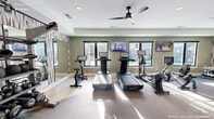 Located on TW Alexander Drive in Durham, NC, this fitness center offers a thoughtfully designed single-floor exercise space with everything needed for a comprehensive workout. The main exercise room is bright and welcoming, filled with abundant natural light streaming through multiple black-framed windows that frame views of an outdoor pool area. Soft sage-green walls, white trim, and a neutral-toned flooring create a clean, modern aesthetic throughout.

The cardio zone features a lineup of treadmills, stationary bikes, and an elliptical machine, each equipped with individual digital screens for interactive workouts. Two wall-mounted flat-screen televisions are positioned for easy viewing from the cardio equipment. Overhead, ceiling fans with sleek, dark-bladed designs provide airflow and comfort during intense sessions.

The strength training area includes a multi-station cable machine and a well-stocked dumbbell rack, complemented by large floor-to-ceiling mirrors accented with bold geometric-patterned wallcovering. The mirrors amplify the sense of space and allow users to monitor their form. Recessed lighting supplements the natural light, ensuring the room remains well-lit at all times.

A connecting hallway provides access to the fitness center's entrance, completing this efficient and stylish community fitness facility.