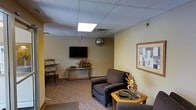 This single-floor space presents a well-appointed waiting or reception area designed with functionality and comfort in mind. The room features a drop tile ceiling with recessed panel lighting that provides even, neutral illumination throughout the space. Warm yellow-toned walls create an inviting atmosphere, complemented by patterned carpeting that adds texture underfoot.

Seating is thoughtfully arranged to accommodate visitors, with a plush upholstered sofa and coordinating armchairs offering a comfortable place to relax. A glass-topped side table with a wooden frame serves as a functional accent piece between the seats. Against the far wall, a wooden console table holds a decorative floral arrangement, adding a touch of warmth and color to the room.

A wall-mounted flat-screen television is positioned for easy viewing from the seating area, enhancing the visitor experience. Framed artwork on the walls contributes to the professional yet welcoming character of the space. A round wall clock and a security dome camera are also visible, reflecting the practical nature of the environment. Overall, the space is designed to be clean, organized, and comfortable for guests awaiting service.