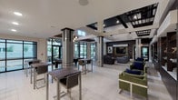 This expansive clubroom is designed to accommodate a variety of social activities within a single, well-appointed space. The architecture draws immediate attention with its soaring ceilings, dramatic structural columns with dark trim accents, and an open-concept layout that connects multiple functional zones seamlessly.

The lounge area anchors the space with generous sectional sofas in warm gray tones, accented by plaid and navy throw pillows arranged around a reflective glass coffee table. A wall-mounted flat-screen television is set against rich dark wood paneling, accompanied by bold abstract artwork. Illuminated display shelving with decorative objects adds a curated, gallery-like quality to this area.

A gaming and casual dining zone offers bar-height tables and chairs alongside a billiards table, positioned near floor-to-ceiling windows that invite abundant natural light and views of the outdoor surroundings.

The fully equipped kitchen features crisp white upper cabinetry, dark lower cabinets, white quartz countertops, a textured tile backsplash, and stainless steel appliances including a range, dishwasher, and microwave. The space is finished with large-format light tile flooring that continues throughout, unifying the entire clubroom with a clean, contemporary aesthetic.