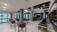 This expansive clubroom is designed to accommodate a variety of social activities within a single, well-appointed space. The architecture draws immediate attention with its soaring ceilings, dramatic structural columns with dark trim accents, and an open-concept layout that connects multiple functional zones seamlessly.

The lounge area anchors the space with generous sectional sofas in warm gray tones, accented by plaid and navy throw pillows arranged around a reflective glass coffee table. A wall-mounted flat-screen television is set against rich dark wood paneling, accompanied by bold abstract artwork. Illuminated display shelving with decorative objects adds a curated, gallery-like quality to this area.

A gaming and casual dining zone offers bar-height tables and chairs alongside a billiards table, positioned near floor-to-ceiling windows that invite abundant natural light and views of the outdoor surroundings.

The fully equipped kitchen features crisp white upper cabinetry, dark lower cabinets, white quartz countertops, a textured tile backsplash, and stainless steel appliances including a range, dishwasher, and microwave. The space is finished with large-format light tile flooring that continues throughout, unifying the entire clubroom with a clean, contemporary aesthetic.
