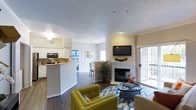 Located at 6805 West Bowles Avenue in Littleton, Colorado, this single-floor residence offers a thoughtfully designed open-concept layout that promotes a comfortable and functional living experience. The living area serves as the heart of the home, highlighted by a classic white-mantled fireplace, a wall-mounted television, and sliding glass doors that welcome abundant natural light and provide direct outdoor access. Rich, dark hardwood-style flooring runs throughout, adding warmth and visual continuity to the space. The kitchen is positioned adjacent to the living area and features white cabinetry, stainless steel appliances including a microwave and range, and a breakfast bar with a light-toned countertop that creates a natural divide between the cooking and living spaces. A pendant light fixture adds a touch of character above the dining area, which is visible just beyond the kitchen. The open sightlines connect each area seamlessly, creating a sense of spaciousness throughout. Textured ceilings and neutral wall tones provide a clean, versatile backdrop, while large windows ensure the interior remains bright throughout the day. The overall ambiance is modern yet approachable, blending practical design with inviting aesthetic details suited for everyday living.