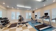 This fitness center offers a comprehensive workout environment designed to accommodate a wide range of exercise needs. The space is finished with a distinctive two-tone checkered tile floor in tan and blue, creating clearly defined zones for different types of training. Large windows fitted with white horizontal blinds line the walls, allowing ample natural light to fill the room while providing views of the surrounding greenery.

The cardio section is well-appointed with treadmills, an elliptical trainer, and a recumbent stationary bike, each equipped with individual monitors for entertainment or performance tracking. A wall-mounted television adds to the amenity.

The strength training area features a Precor functional trainer cable machine, ideal for a wide variety of resistance exercises, along with a weight bench for targeted workouts. A full rack of dumbbells in graduated sizes is neatly organized in a dedicated corner, complemented by a large floor-to-ceiling mirror that enhances the sense of space and supports proper form.

Rubber floor mats are positioned throughout for floor exercises and stretching. Recessed and overhead fluorescent lighting ensures the space remains bright at all hours. The fitness center is accessible via an adjoining hallway, making it a convenient and functional facility.