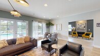 This well-appointed single-floor community clubhouse is designed to offer residents a comfortable and functional gathering space. The main lounge area is anchored by light hardwood flooring, sage green and charcoal accent walls, crown molding, and recessed lighting throughout. Large windows and French doors flood the space with natural light, offering direct views of and access to the outdoor pool and patio area. The lounge is furnished with a mix of leather and upholstered seating arranged around layered area rugs, creating distinct yet open zones for relaxation and socializing. A sculptural multi-arm pendant light adds a modern focal point overhead. Adjacent to the lounge, a dedicated dining nook features a round table with upholstered chairs, framed wall art, and a tall fiddle-leaf fig plant, lending a warm, residential feel. A secondary lounge area is highlighted by a floor-to-ceiling tile fireplace surround, bold dark walls, and a striking abstract canvas above the mantle, complemented by a vibrant yellow accent chair. The kitchenette features dark cabinetry, stainless steel appliances, and a bar-height counter with wood stools. A dedicated business center offers a built-in desk with dual computer workstations, printing equipment, and a live plant wall panel, making it ideal for remote work needs.