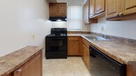 This single-story unit offers a well-organized layout with multiple rooms spread across a thoughtfully arranged floor plan. The interior maintains a consistent, neutral color palette of warm cream and beige tones throughout, creating a cohesive and understated atmosphere.

The living area is generously sized, featuring plush wall-to-wall carpeting and multiple windows fitted with blinds that allow ample natural light to fill the space. Several additional carpeted rooms serve as bedrooms, each with windows providing natural light. At least one bedroom includes a standard closet with a rod and shelf, while another features a double-door closet, offering practical storage solutions.

The kitchen is outfitted with warm wood-toned cabinetry, granite-look countertops, and a full suite of black appliances including an electric range with overhead ventilation, a dishwasher, and a refrigerator. Light ceramic tile flooring ties the kitchen together. A dedicated laundry area with stacked washer and dryer units adds everyday convenience.

Overall, the unit presents a clean, functional layout with consistent finishes, practical storage, and well-distributed natural light throughout its rooms.