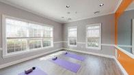 This single-floor wellness studio is thoughtfully designed to support yoga and mindfulness practices in a calm, functional environment. The main practice room is the heart of the space, finished with light wood-grain flooring and soft gray walls accented by crisp white crown molding and chair rail trim. Multiple large windows with horizontal blinds line the walls, flooding the room with natural light and offering views of surrounding greenery. Purple yoga mats and foam blocks are arranged across the open floor, providing ample room for movement and stretching. Recessed ceiling lighting supplements natural illumination, ensuring a well-lit environment at any hour.

A bold accent wall painted in vibrant orange adds energy to one side of the studio, featuring a large wall-mounted mirror that enhances the sense of space and allows practitioners to monitor their form. A mounted screen is also visible, supporting guided or virtual instruction. Adjacent to the practice area, a built-in cabinetry station features light blue lower cabinets with brushed nickel hardware topped by a sleek, dark granite-style countertop, offering practical storage and surface space. The overall ambiance is bright, open, and serene, balancing energizing design elements with a tranquil atmosphere suited for focused wellness activities.