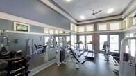 This single-floor fitness center offers a thoughtfully designed workout environment suited for a range of exercise routines. The space features a generous ceiling height accentuated by crown molding and a two-toned wall treatment in soft blue-gray and cream, lending the room a clean, polished aesthetic. A ceiling fan provides additional air circulation throughout the space.

Large windows and glass doors line the perimeter, allowing abundant natural light to flood the interior and providing views of the surrounding exterior. Full-length mirrors along one wall visually expand the space and serve the practical needs of those monitoring their form during strength training.

The fitness center is equipped with a variety of strength machines, including a multi-function cable and resistance unit, as well as multiple treadmills to accommodate cardio training. Additional exercise equipment is positioned throughout the room, offering users a well-rounded selection of workout options. The flooring is a durable, rubberized surface appropriate for a high-activity fitness environment.

Overall, the space strikes a balance between functionality and comfort, with its bright, airy atmosphere and comprehensive equipment selection making it a practical and inviting facility for daily exercise.