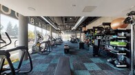 Located at 18401 Nordhoff St in the Northridge neighborhood of Los Angeles, this single-floor fitness center offers a spacious, well-equipped workout environment with a contemporary industrial aesthetic. The facility features exposed ductwork and piping along the ceilings, teal and charcoal patterned carpet tiles throughout, and floor-to-ceiling windows along the perimeter that flood the space with natural light and provide open views of the surrounding streetscape.

The cardio zone is lined with a wide selection of treadmills, ellipticals, stair climbers, and stationary bikes — all arranged to take advantage of the expansive window views. A dedicated strength training area houses multiple racks of dumbbells, weight benches, and Hoist cable machines, providing a comprehensive free weights section. Additional functional training equipment, including medicine balls, kettlebells, and plyo boxes, is neatly organized on wall-mounted shelving units.

A lounge area with seating and a bold, geometric multicolor mural adds a vibrant focal point to one section of the facility, while wall-mounted TVs are distributed throughout. Branded signage and a colorful, energetic design language give the space a distinct, motivating atmosphere well-suited for an active fitness community.