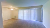 This single-floor space centers around a generously sized living area that offers a clean, neutral aesthetic. The room is finished with wall-to-wall carpeting in a warm beige tone, providing a comfortable and cohesive foundation throughout the space. A prominent feature of the living area is the expansive sliding glass door spanning nearly the full width of one wall, dressed with white vertical blinds that allow for flexible light control while maximizing the sense of openness. Natural light filters in around the edges of the blinds, brightening the room with a soft, diffused glow. The walls are painted in a light, neutral tone that complements the overall brightness of the space. A ceiling fan with an integrated light fixture is centrally positioned overhead, offering both air circulation and ambient lighting. A floor-level vent is visible along the exterior wall, indicating a built-in climate control system. The room's layout is open and versatile, providing ample space for a variety of furniture arrangements. The overall ambiance is clean, bright, and move-in ready, offering a functional and comfortable living environment.