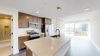 This single-floor residence showcases a thoughtfully designed open layout that seamlessly connects the kitchen and living areas. The kitchen serves as the centerpiece of the space, anchored by a generously sized waterfall quartz island equipped with an undermount sink and matte black faucet. Rich dark-toned wood cabinetry with gold hardware lines the walls, complemented by a white subway tile backsplash and a full suite of stainless steel appliances, including a gas range, built-in microwave, and French door refrigerator. Gold-accented pendant and linear lighting fixtures add a refined touch overhead.

The living area benefits from abundant natural light pouring through floor-to-ceiling windows and a glass-panel door, framing striking views of a waterway and the surrounding cityscape. An exposed concrete accent wall adds an industrial architectural element, contrasting with the light wood-tone flooring that runs throughout the main living spaces.

The bathroom features large-format tile work, a frameless glass-enclosed walk-in shower with matte black hardware, and a built-in open shelving unit for organized storage. Additional rooms offer neutral wall tones and versatile flooring, providing a clean, move-in-ready canvas throughout the residence.
