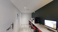 This single-floor business center is designed with both functionality and modern aesthetics in mind. The room features a long, continuous terrazzo-style countertop workstation running along one wall, equipped with multiple desktop computers and a multifunction printer, providing ample workspace for several users simultaneously. Vibrant red ergonomic chairs offer comfortable seating at each station, adding a striking pop of color against the neutral surroundings.

The design is defined by a compelling contrast between a crisp white painted brick accent wall and a deep forest green feature wall, creating a dynamic visual atmosphere. Inspirational typographic artwork, including phrases like "WHY?" and "BELIEVE," is displayed on the white brick wall, adding a motivational character to the space.

Recessed ceiling lighting provides consistent, focused illumination throughout the room. Light-toned, patterned carpet tiles cover the floor, contributing to the overall clean and contemporary feel. A dark-framed doorway leads to an adjoining corridor, offering convenient access to surrounding areas. The space is well-suited for productivity and professional use.