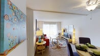 This single-floor residence features a cozy and vibrantly decorated living area that serves as the focal point of the space. The room is anchored by a large sliding glass door or wide window dressed in sheer white curtains, allowing abundant natural light to pour in and creating a bright, airy atmosphere. The living area is generously sized, accommodating a comfortable seating arrangement that includes a bold red sofa and accent chairs, complemented by a stylish, patterned area rug with colorful circular motifs that add energy and personality to the space. A low-profile media console houses a flat-screen television and is flanked by tall, slender floor lamps that provide warm, ambient lighting. Decorative wall art and framed prints contribute to the space's cheerful aesthetic. A small side table with a gourd-style lamp adds a charming, eclectic touch near the room's entrance. The white walls and smooth ceilings maintain a clean, neutral backdrop that allows the colorful furnishings to stand out. Overall, the space presents a warm, inviting ambiance with a well-curated blend of color, texture, and light.
