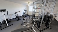 Located at 38241 Lakeshore Blvd. in Willoughby, Ohio, this fitness center is a well-equipped, single-floor exercise facility designed to support a well-rounded workout routine. The room features durable dark rubber flooring suited for heavy equipment use, and light-colored walls that keep the space feeling open and clean. A ceiling fan provides air circulation throughout the room. Natural light filters in through a window dressed with vertical blinds, adding a bright, welcoming atmosphere to the workout environment. At the heart of the space sits a large multi-station cable and weight machine, offering a wide range of strength training exercises. Complementing it is a dedicated bench press setup complete with a barbell and weight plates. A spin bike positioned along the wall provides a solid option for low-impact cardiovascular training, while a motorized treadmill serves those looking for walking or running workouts. Additional free weights and accessories are accessible throughout the room. The layout is efficiently arranged, allowing users to move between equipment with ease. Overall, this fitness center provides a practical and functional space for both cardio and strength training without leaving the property.