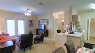 Located at 10900 East Taylor Road in Gulfport, Mississippi, this single-story residence offers a thoughtfully designed open-concept floor plan that promotes a seamless flow between living and functional spaces. The living area is comfortably proportioned, featuring plush carpeted flooring, neutral-toned walls, and crown molding that adds a refined architectural touch. A ceiling fan with a light fixture serves as a practical centerpiece overhead, while a large window allows natural light to brighten the room. The living space is well-suited for relaxed seating arrangements, centered around an entertainment console. Adjacent to the living area, a raised counter or breakfast bar creates a natural transition into the kitchen and dining zone, providing additional workspace and casual seating opportunities. A desk area near the counter offers a dedicated spot for work or study. A hallway visible from the main living space leads to additional rooms, suggesting a well-organized layout that keeps private areas separate from shared living zones. Throughout the space, the consistent use of light-toned finishes, carpeted floors, and warm lighting contributes to a cohesive, inviting ambiance that is both functional and comfortable.