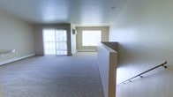 This single-floor space features an open, airy layout highlighted by abundant natural light streaming through multiple large windows and a full sliding glass door. The sliding door leads to an exterior area, allowing light to flood the main living space throughout the day. Neutral-toned walls complement the plush wall-to-wall carpeting underfoot, creating a clean, cohesive aesthetic throughout. A baseboard heating unit runs along the far wall beneath the windows, providing efficient climate control. A partial half-wall with a staircase railing is visible to one side, defining the entry point into the main living area while maintaining an open feel. A wall-mounted unit, likely an air conditioner or ventilation fixture, is positioned near the sliding door, adding to the functional comfort of the space. The smooth, light-colored ceiling features a recessed light fixture, contributing to the clean and uncluttered atmosphere. Overall, the space presents a straightforward, functional layout with ample room for furnishing, benefiting from natural light and a simple, neutral palette that offers versatility for a variety of interior arrangements.
