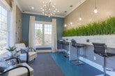 Located at 101 Peaks Landing in Acworth, Georgia, this single-floor space is designed with a contemporary aesthetic that balances function and style. The interior is defined by a cohesive color palette of teal, gray, and warm taupe, creating a polished and inviting atmosphere throughout.

The main area features a sleek workstation zone with built-in desks finished in crisp white, complemented by textured slate-toned accent walls and lush green ornamental grass planters that add a natural, refreshing element. Modern globe pendant lights hang above the workspace, casting a warm, ambient glow. Adjustable chrome bar-height chairs offer ergonomic seating at the computer stations.

A distinct seating lounge area offers comfortable white upholstered chairs with blue accent pillows, positioned alongside large windows dressed with sheer white curtains that allow generous natural light and outdoor views of the surrounding neighborhood. Crown molding frames the ceiling throughout, adding an architectural refinement.

An arched architectural detail elegantly divides the space, creating a sense of definition between zones while maintaining an open, connected feel. The flooring transitions between plush gray carpet and vibrant teal hardwood-style planks, reinforcing the space's bold, modern character. A glass-panel entry door provides additional natural light near the workstation area.