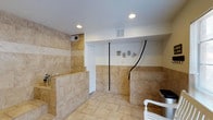 This purpose-built pet grooming facility is thoughtfully designed to provide a clean, functional, and welcoming environment for dogs and their owners. The space is finished with warm, beige ceramic floor and wall tiles that extend halfway up the walls, creating a durable and easy-to-clean surface throughout. Two dedicated dog wash stations are the centerpiece of the room, each enclosed by tiled half-walls and equipped with handheld spray attachments, offering ample space for bathing pets of various sizes. Recessed lighting in the white ceiling provides bright, even illumination across the entire space, ensuring groomers have excellent visibility. Ventilation is addressed with wall and ceiling HVAC vents, keeping the environment comfortable for both animals and staff. A playful "WOOF" sign mounted on the white upper wall adds a charming, dog-friendly character to the space. Black grooming tethers are conveniently anchored to the wall between the stations, keeping pets safely secured during the grooming process. A white storage unit is partially visible near the entrance, offering practical organization for grooming supplies. The single-floor layout ensures easy accessibility and a seamless workflow throughout the grooming process.