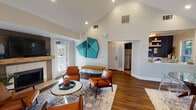 This thoughtfully designed single-story space makes an immediate impression with its dramatic vaulted ceilings and crisp white walls, creating an open, airy atmosphere throughout. The main living area serves as the focal point, anchored by a wood-framed fireplace with a tile surround and a wall-mounted television above. Mid-century modern cognac leather chairs arranged around a round marble-top coffee table establish a refined, welcoming seating arrangement, complemented by a patterned area rug over rich hardwood flooring.

Bold teal geometric wall art adds a striking visual element, echoing the color accents found throughout the space. Recessed lighting and a circular chandelier provide warm illumination, while multiple windows and glass doors invite abundant natural light. Elegant arched doorways connect the main area to additional rooms, including a workspace furnished with a desk and seating, and a staircase landing bathed in natural light.

The galley-style kitchenette features light cabinetry with white countertops, a decorative navy patterned accent wall, open shelving, a microwave, and a sink with ample prep space. Tile flooring and a window overlooking the outdoors complete the functional yet stylish kitchen area.