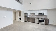 This two-story loft showcases a dramatic, open-concept layout defined by soaring double-height ceilings that create an expansive, airy atmosphere. The upper level, accessible via an interior overlook with a metal railing, adds architectural depth and versatile living space.

The main floor features polished concrete flooring throughout, lending the space an industrial yet contemporary character. The kitchen is well-appointed with white upper cabinetry contrasted by warm wood-toned lower cabinets, a subway tile backsplash, and a suite of stainless steel appliances including a refrigerator, electric range, built-in microwave, and dishwasher. A functional single-basin sink with a modern faucet completes the setup.

Adjacent to the kitchen, a dedicated laundry alcove houses a side-by-side washer and dryer behind louvered bi-fold doors, offering convenient in-unit laundry while maintaining a clean, organized aesthetic. The white walls and high ceilings amplify natural light, contributing to the bright, modern ambiance that defines the overall character of this multi-level space.