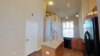 This two-story residence offers three bedrooms and two bathrooms across a well-designed floor plan. The main living area immediately impresses with its soaring double-height ceilings, creating an expansive, airy atmosphere that draws the eye upward to the open upper level above.

The kitchen is a central highlight of the main floor, featuring warm wood cabinetry, granite countertops, and a center island with pendant lighting overhead. A built-in dishwasher and sink with a window view add both functionality and natural light to the space. Track lighting on the ceiling complements the pendant fixtures, ensuring the kitchen is well-lit throughout.

A wooden staircase with light-toned railings connects the main floor to the upper level, where an open loft-style landing overlooks the living area below, equipped with a ceiling fan. This upper level adds architectural dimension while providing additional functional space.

The main floor also benefits from a large arched window that floods the living area with natural light. Durable hardwood-style flooring runs throughout, tying the spaces together cohesively and adding warmth to the overall design.
