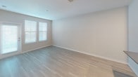 This single-floor residence is designed with a clean, contemporary aesthetic throughout. The open-concept layout connects the main living area to the kitchen, both finished with light-toned hardwood flooring that contributes to a bright, airy feel. Large windows fitted with horizontal blinds allow natural light to filter into the living spaces.

The kitchen is well-appointed, featuring white shaker-style cabinetry extending to the ceiling, gray countertops, a tiled backsplash, and a full suite of stainless steel appliances, including a refrigerator, electric range, and over-the-range microwave. A peninsula provides additional counter space and casual seating potential.

The bedroom is carpeted in a neutral tone and receives natural light through a pair of windows. An additional room, accessible from the main living area, is also carpeted and similarly understated in its finishes.

The bathroom is notably elongated, featuring light wood-look tile flooring, white cabinetry, a dark stone-topped vanity with an undermount sink, an illuminated mirror, a walk-in shower with glass enclosure, and a separate toilet area. The neutral color palette and clean lines carried throughout the space lend it a cohesive, modern character.