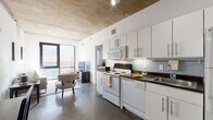 This single-floor urban unit showcases a distinctive industrial-chic design, defined by exposed concrete ceilings with visible structural elements and polished concrete floors that run seamlessly throughout the space. The open layout connects key living areas with a cohesive, contemporary feel.

The kitchen is well-appointed with white cabinetry featuring sleek bar-pull hardware, dark countertops, a full suite of white appliances including a refrigerator, electric range, built-in microwave, and dishwasher. A stainless steel sink and under-cabinet lighting complete the functional and clean aesthetic.

The living area benefits from large floor-to-ceiling windows that frame views to the exterior and flood the room with natural light. The space is versatile, accommodating both a seating area and a dedicated desk workspace, making it suitable for both relaxing and remote work.

The bedroom continues the industrial theme with exposed concrete accent walls alongside softer painted surfaces, a window with blinds, and warm hardwood-style flooring that adds a cozy contrast to the raw architectural elements.

The bathroom is clean and minimalist, featuring a white vanity with an integrated sink, a large mirror, and polished white fixtures. Throughout the unit, the interplay of raw concrete, white walls, and modern finishes creates a sophisticated urban ambiance.
