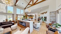 Located at 10015 N Owasso Expressway in Owasso, Oklahoma, this single-floor community clubhouse offers a generous and thoughtfully designed gathering space. The architecture immediately impresses with dramatic vaulted ceilings accented by exposed wood beam trusses, flooding the interior with a sense of grandeur and warmth. Oversized windows and glass doors line the walls, inviting abundant natural light and offering views of the surrounding outdoor areas.

The open living room serves as the heart of the space, anchored by a sleek linear electric fireplace set within a fluted wood surround topped with a marble surface. Elegant pendant chandeliers and light hardwood flooring flow throughout, creating a cohesive and inviting atmosphere. A dedicated lounge area near the entrance features soaring clerestory windows and a sculptural orb chandelier.

The fully equipped kitchen is a standout feature, showcasing floor-to-ceiling natural wood cabinetry, a lengthy marble-topped island with bar seating, bold gold pendant lighting, stainless steel appliances, and a marble backsplash. Adjacent to the kitchen, a billiards room with vibrant patterned flooring and ample natural light provides a recreational retreat. A workstation area with built-in desks rounds out this well-appointed communal space.