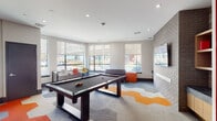 This contemporary amenity lounge is thoughtfully designed to blend recreation and relaxation into a single, cohesive space. The room makes an immediate impression with its bold, geometric-patterned carpet in gray, white, and orange, which anchors the entire layout and adds a vibrant energy to the interior.

At the center of the space sits a full-sized pool table, serving as the focal point of the recreational area. Surrounding it, a variety of seating arrangements cater to different activities — from a sleek leather sofa paired with colorful accent ottomans, to casual dining-height tables and chairs in bright orange and yellow hues positioned near the windows.

One wall features exposed brick detailing with built-in wooden shelving, a wall-mounted flat-screen television, and a cushioned built-in bench, creating a cozy media nook ideal for lounging. On the opposite side, horizontal wood-plank wall cladding adds warmth and a natural texture that contrasts beautifully with the modern, clean-lined architecture throughout.

Large floor-to-ceiling windows line multiple walls, flooding the space with natural light and offering open views of the surrounding streetscape. The entry area features glass sliding doors, bold orange accent walls, and large-format tile flooring, setting a striking and modern tone from the moment of arrival. Recessed lighting throughout ensures the space remains bright and inviting at all hours.