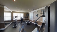 This single-floor fitness space is thoughtfully designed to accommodate a comprehensive workout experience. The room features a dark, durable flooring surface well-suited for exercise equipment, complemented by warm-toned walls accented with crisp white crown molding and baseboards that lend a polished, finished look to the interior.

Recessed lighting is evenly distributed across the ceiling, ensuring the space is well-lit at all times. Multiple windows fitted with white horizontal blinds line the walls, allowing an abundance of natural light to fill the room while maintaining privacy.

The fitness area is outfitted with a broad selection of equipment, including an elliptical trainer, a stationary recumbent bike, a treadmill, and a multi-station cable strength machine, providing options for both cardiovascular conditioning and resistance training. A wall-mounted television is positioned for easy viewing during workouts, adding a convenient entertainment feature.

The layout is efficiently organized, allowing users to navigate between machines with ease. The combination of functional equipment, thoughtful design details, and a bright, open atmosphere makes this fitness room a practical and inviting space for daily exercise.