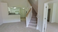 This two-story space offers a functional and straightforward layout designed for comfortable everyday living. The main level features wall-to-wall carpeting in a neutral tone that extends throughout the open living area, creating a cohesive and understated aesthetic. The staircase, positioned centrally within the space, is finished with matching carpet treads and a warm wood handrail supported by clean white balusters, providing a classic and practical connection between floors.

Adjacent to the living area, the kitchen is visible from the main living space, offering an open feel between the two areas. The kitchen features white cabinetry, a dark countertop, and under-cabinet lighting, providing a clean, functional workspace. A standard entry door is positioned nearby, contributing to the flow of the main level.

The white walls and ceilings throughout maintain a bright, neutral atmosphere, while flush-mounted ceiling light fixtures provide consistent illumination across the space. The overall design prioritizes practicality, with a clean, move-in-ready presentation across both levels.