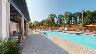 The outdoor amenity area at Pointe Grand Apartment Homes is designed to deliver a resort-style experience for residents. At the heart of the space is a generously sized swimming pool with crystal-clear blue water, complemented by a separate shallow wading area, making it suitable for a range of recreational activities. The pool deck is finished with attractive paver stonework that extends throughout the entire outdoor area, providing a polished and cohesive aesthetic.

Surrounding the pool, multiple lounge chairs and seating areas are arranged to take full advantage of the open sky and natural sunlight. Yellow market umbrellas provide shaded retreats along the deck, while a natural wood storage structure adds a functional and visually warm accent to the space.

Adjacent to the pool, a covered cabana lounge area offers a more private and sheltered setting. This structure features a dark-paneled ceiling with ceiling fans, white board-and-batten walls, and elegant floor-to-ceiling drape curtains that can be drawn for privacy. The cabana is furnished with comfortable seating and is decorated with tropical potted plants and colorful accents, creating a welcoming, leisure-focused environment. Double glass entry doors bearing the Pointe Grand branding connect the cabana to the pool deck. A mature tree line and landscaped hillside form a natural backdrop to the entire area.