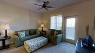This single-floor space offers a warm and inviting atmosphere centered around a well-appointed living room. The living area is furnished with a spacious green sofa and matching loveseat, complemented by a patterned ottoman that doubles as a functional centerpiece. Soft carpeting underfoot adds to the room's comfort and casual charm.

Natural light filters in through vertical blinds covering a large window, while a glass-paneled door provides additional brightness and a glimpse of the greenery outside. A ceiling fan with wooden blades is mounted overhead, promoting airflow and contributing to the room's relaxed, functional character.

Warm ambient lighting from a classic table lamp enhances the cozy feel of the space during evening hours. The neutral wall tones create a versatile backdrop that complements the earthy green tones of the furnishings and the accent patterns of the decorative pillows and ottoman.

Overall, the space presents a functional and comfortable living environment, with thoughtful touches that balance practicality and homeliness. The layout encourages easy movement and social interaction, making it well-suited for everyday living.