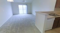 This single-floor space offers a clean, open layout designed for comfortable living. The main living area is generously sized, featuring plush wall-to-wall carpeting that adds warmth underfoot. A ceiling fan overhead provides both functionality and a subtle design touch, while the room's neutral white walls create a bright, airy atmosphere throughout.

Natural light pours in through a large sliding glass door, which opens to a private balcony enclosed by a classic railing. The balcony offers views of the surrounding exterior, making it an inviting spot to enjoy the outdoors. The open sightlines from the living area to the balcony enhance the sense of space and airflow.

Adjacent to the living area, a partial wall or peninsula element — likely part of the kitchen — defines the boundary between spaces while maintaining an open-concept feel. The light-toned countertop surface hints at a functional and neatly appointed kitchen nearby.

Overall, the space is characterized by its clean lines, neutral palette, and abundance of natural light. The combination of soft carpeting, open layout, and indoor-outdoor connectivity creates a welcoming and versatile environment well-suited for everyday living.