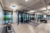 Located at 2651 156th Ave NE in Redmond, Washington, this expansive fitness facility offers a generously proportioned, open-plan layout spanning a single floor. The space features an industrial-modern design aesthetic, with exposed ceiling ductwork and structural concrete columns that lend a clean, loft-like character throughout. Light wood-plank flooring runs continuously across the entire area, contributing to a bright, cohesive atmosphere complemented by abundant linear LED lighting overhead.

The facility is organized into distinct functional zones. A dedicated strength training area houses a multi-station cable machine, power racks, and a full dumbbell rack accompanied by rubber floor mats for safe lifting. A separate cardio zone lines the perimeter near large windows, equipped with treadmills and other aerobic machines that benefit from natural light and outdoor views. An open floor zone provides ample space for functional training, featuring tire sleds and additional freestanding equipment.

A stylishly appointed lounge area near the entrance features floor-to-ceiling glass partitions, sheer drapery, and contemporary furnishings, offering a welcoming transition into the fitness space. A glass-railed mezzanine overlook connects to an adjacent lower-level lounge, adding vertical dimension and architectural interest. The overall ambiance is polished yet functional, designed to accommodate a wide range of fitness activities.