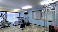 This fitness center offers a focused and functional workout environment spread across a single level. The room is finished with cool blue walls and a neutral patterned carpet that adds texture underfoot. Bright flush-mounted ceiling lights ensure the space is well-lit throughout the day, complemented by natural light streaming in through multiple windows fitted with roller shades, which also provide views of the surrounding greenery.

The cardio section is well-appointed, featuring two treadmills positioned along the windowed wall and a recumbent stationary bike, catering to a range of aerobic exercise preferences. A wall-mounted flat-screen television is positioned for easy viewing during workouts. On the opposite side of the room, a multi-function cable machine provides options for strength and resistance training.

The layout is practical and open, allowing users to move between equipment comfortably without feeling confined. The overall ambiance is clean and utilitarian, designed with functionality in mind. This fitness center provides the essential amenities needed for a balanced exercise routine, making it a convenient and accessible facility for daily use.