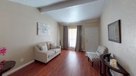 This single-floor residence combines functional design with comfortable living across several well-defined spaces. The living room immediately draws attention with its vaulted, beam-accented ceiling and rich hardwood flooring, creating an open and airy atmosphere. Natural light filters through grommet-panel curtains framing the front entry, while crown molding adds a refined architectural detail consistent throughout the home.

The kitchen features white cabinetry, granite-look countertops, a double stainless steel sink, and tile flooring, opening directly to a combined dining and laundry area. This multifunctional space accommodates a round dining table with seating for four alongside a full-size washer and dryer, all finished with crown molding and a ceiling light fixture.

The primary bedroom offers carpeted flooring, crown molding, a ceiling fan, a window with drape panels, and access to a walk-in closet. A second carpeted bedroom mirrors this comfortable feel with similar finishes. A flexible bonus room with carpet provides additional utility as a home office or sitting area. The bathroom presents a vanity with granite-look countertop and wall sconce lighting. Ceiling fans are present throughout, contributing to a consistently comfortable and cohesive interior.