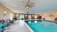 Located at 445 Warfield Blvd in Clarksville, Tennessee, this generously sized indoor aquatic facility offers a resort-style experience within a thoughtfully designed single-story structure. The centerpiece is a large, sparkling indoor swimming pool with clearly marked depth indicators, framed by a wide, stamped concrete pool deck that provides ample space for lounging and recreation. A separate hot tub area is also present, adding to the facility's leisure offerings. Multiple large ceiling fans are mounted overhead, ensuring comfortable air circulation throughout the space, complemented by recessed lighting that keeps the interior bright and inviting. The pool deck is lined with classic Adirondack chairs paired with colorful accent side tables, creating a relaxed, welcoming atmosphere. Numerous windows along the perimeter walls allow generous natural light to filter in while offering views to the outdoors. The walls feature a warm two-tone color scheme of soft gray and beige, accented by decorative wall-mounted sconces. A basketball hoop and a water slide add recreational variety. Double doors and multiple entry points provide convenient access throughout the facility, while safety equipment and amenities such as a drinking fountain are thoughtfully integrated into the space.