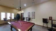 This single-floor recreation space is thoughtfully designed for social activity and relaxed entertainment. At the heart of the room sits a full-sized pool table with a rich burgundy felt surface and a classic wood frame, positioned beneath a striking Tiffany-style pendant light fixture suspended by chains — a standout decorative element that adds warmth and character to the space.

The room features neutral, cream-toned walls that create a bright and welcoming atmosphere, complemented by recessed lighting and carpeted flooring that enhances the overall comfort of the area. Natural light filters in through a window on one side, adding to the open, airy feel of the space.

Along the walls, framed decorative prints add visual interest, while a small seating area with wicker-style chairs and a café table provides a spot for conversation or casual games. A tall indoor plant near the window introduces a natural element, softening the room's décor. Additional bar-height seating is also present, rounding out the versatile social layout of this inviting recreation room.