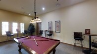 This single-floor recreation space is thoughtfully designed for social activity and relaxed entertainment. At the heart of the room sits a full-sized pool table with a rich burgundy felt surface and a classic wood frame, positioned beneath a striking Tiffany-style pendant light fixture suspended by chains — a standout decorative element that adds warmth and character to the space.

The room features neutral, cream-toned walls that create a bright and welcoming atmosphere, complemented by recessed lighting and carpeted flooring that enhances the overall comfort of the area. Natural light filters in through a window on one side, adding to the open, airy feel of the space.

Along the walls, framed decorative prints add visual interest, while a small seating area with wicker-style chairs and a café table provides a spot for conversation or casual games. A tall indoor plant near the window introduces a natural element, softening the room's décor. Additional bar-height seating is also present, rounding out the versatile social layout of this inviting recreation room.