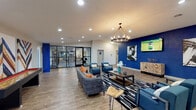 This single-floor amenity space is thoughtfully designed to offer residents a variety of social, recreational, and functional areas within one cohesive layout. The central lounge serves as the heart of the space, featuring wood-look flooring, a bold cobalt blue accent wall, and a striking circular chandelier that anchors the room with an air of modern sophistication. Comfortable seating — including a plush gray sofa with orange accent pillows and coordinating blue armchairs — is arranged around a black-and-white geometric area rug, creating an inviting gathering area. Contemporary artwork adorns the walls, and a wall-mounted flat-screen TV adds an entertainment focal point. Recessed lighting throughout ensures a well-lit, welcoming atmosphere. Adjacent to the lounge, a glass-walled fitness center offers full visibility into a well-equipped gym, providing an open, connected feel between the activity areas. A game area with a pool table adds a recreational element, while a dedicated computer workstation nook provides a practical space for productivity. The layout also includes a hallway corridor leading to additional rooms and restroom facilities. Together, these areas create a versatile, amenity-rich environment designed to foster community and comfort.