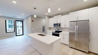 Located in Bloomington, Indiana, this contemporary 3-bedroom, 1-bathroom unit is thoughtfully designed with a clean, modern aesthetic across a single floor. The open-concept kitchen and living area serves as the heart of the home, showcasing crisp white shaker cabinetry, a patterned tile backsplash, and a full suite of stainless steel appliances including a refrigerator, electric range, and built-in microwave. A large center island with a white quartz countertop and undermount sink anchors the space, complemented by stylish pendant lighting overhead. Recessed lighting and light wood-style flooring flow throughout, creating a bright and cohesive atmosphere. The three bedrooms feature neutral gray walls and black-framed windows that invite natural light and offer views of the surrounding neighborhood. One bedroom includes a sliding closet, while another provides direct access to a private outdoor balcony through a glass door. The full bathroom is well-appointed with a white vanity, tub-shower combination with white subway tile surround, and brushed nickel fixtures. A dedicated closet offers additional storage. The unit's contemporary finishes and efficient layout make it a functional and stylish living space.