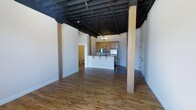 This single-floor loft-style space showcases a distinctive industrial aesthetic with a thoughtfully open layout. The most striking architectural feature is the exposed ceiling with dark-painted structural joists and beams, lending dramatic height and an authentic urban character to the interior. Warm, natural hardwood flooring runs throughout the main living area, providing a welcoming contrast to the raw ceiling above.

The open floor plan flows seamlessly from the expansive main living area toward a kitchen at the rear, which features warm wood cabinetry, stainless steel appliances including a refrigerator and range, and a peninsula-style breakfast bar with overhead shelving — ideal for casual dining or entertaining. A small chandelier above the kitchen adds a touch of elegance to the otherwise industrial setting.

Structural columns are integrated throughout the space, reinforcing the building's original character while subtly defining zones within the open plan. Crisp white walls throughout help reflect light and keep the space feeling bright and airy. Additional rooms are accessible through doorways visible on both sides of the main area, suggesting private spaces such as a bathroom and additional rooms beyond. The overall ambiance is a balanced blend of raw industrial character and livable warmth.