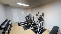 This fitness room offers a dedicated space for cardiovascular exercise, featuring a selection of modern cardio equipment including a treadmill, an elliptical trainer, and a stationary recumbent bike. Each machine is positioned on protective rubber floor mats, preserving the light-toned tile flooring beneath. The room is finished with neutral grey walls and is illuminated by a bright overhead fluorescent panel, ensuring a well-lit environment for workouts at any time of day. A standard white door provides access to the space, and a ventilation grille in the ceiling supports air circulation throughout the room. The overall layout is straightforward and functional, making efficient use of the available floor space to accommodate multiple users. The clean, minimalist design keeps the focus on the equipment, creating a practical and no-frills environment suited for residents or occupants looking for convenient on-site fitness access.