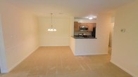 This single-floor residence showcases an open-concept design that seamlessly connects the living, dining, and kitchen areas into one cohesive space. The living and dining area is finished with wall-to-wall carpeting in a neutral beige tone, creating a warm and inviting atmosphere. A chandelier with multiple light fixtures is centrally positioned over the dining area, adding a decorative focal point and providing ambient lighting throughout the space. The walls are finished in a clean, light neutral tone, contributing to a bright and airy feel. The kitchen is defined by a peninsula counter with a dark countertop, which serves as both a workspace and a visual divider between the kitchen and the main living area. Medium-toned wood cabinetry lines the kitchen walls, offering ample storage, while stainless steel appliances — including a microwave and a stove — add a modern touch. A black refrigerator anchors the kitchen, completing the appliance suite. The overall layout emphasizes functionality and flow, making the space well-suited for everyday living and casual entertaining.