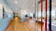This expansive single-floor amenity space offers a thoughtfully designed environment that blends functionality with contemporary style. The open-plan layout features wide-plank hardwood-style flooring throughout, creating a warm and cohesive aesthetic across all areas.

The main lounge area is anchored by floor-to-ceiling windows that frame stunning waterfront views, flooding the space with natural light. Seating arrangements include plush sofas, accent chairs in bold mustard and blue tones, and a variety of flexible furniture configurations suited for both relaxation and social interaction. A striking feature wall adorned with large-scale typographic artwork adds an artistic focal point to one of the lounge zones, while a colorful vertical rod installation serves as a sculptural room divider, injecting personality and visual interest.

A well-appointed kitchenette and service counter provide refreshment amenities, complemented by casual bar-height seating and intimate dining clusters. Private meeting rooms offer a quieter setting, each furnished with round wooden tables, vibrant orange chairs, and curated wall art. Soft, neutral wall tones and statement pendant lighting complete these spaces.

Recessed ceiling lighting, an open corridor connecting the zones, and abundant greenery throughout contribute to an overall ambiance that is bright, energetic, and welcoming.