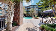 Located at 35 Folly Road Blvd in Charleston, South Carolina, this beautifully designed outdoor courtyard offers a resort-style retreat at the heart of a multi-story residential community. The centerpiece of the space is a sparkling pool, elegantly framed by raised brick planters, swaying Palmetto palms, and manicured tropical greenery that evokes the lush character of the Lowcountry.

Wide, herringbone-patterned brick and paver walkways wind throughout the courtyard, connecting key areas while providing a refined, cohesive aesthetic. Mature trees and dense plantings of ornamental grasses, shrubs, and ground cover create a sense of privacy and natural enclosure, giving the space a garden-like ambiance.

Lounge areas with shaded canopies, including a striped blue-and-white cabana, are positioned throughout the courtyard, offering comfortable outdoor seating and relaxation spots. Wrought iron fencing delineates sections of the space while maintaining an open, airy feel. Ground-level unit entrances with double glass doors open directly onto the courtyard, seamlessly blending indoor and outdoor living.

The multi-story building surrounding the courtyard features balconies that overlook the lush grounds below. Abundant natural light filters through the tree canopy, enhancing the inviting atmosphere of this thoughtfully landscaped outdoor living space.