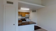 This single-floor residential unit offers a practical and functional layout designed to maximize everyday living. The kitchen is the focal point of the space, featuring warm honey-toned wood cabinetry that provides ample storage both above and below the countertops. A white electric range and oven sit beneath a ventilation hood, accompanied by a sink framed by dark granite-style countertops that add contrast to the lighter surroundings. A peninsula counter with a dark surface extends outward, creating a natural divider between the kitchen and the adjacent living area while offering additional prep or casual dining space. The textured ceiling adds subtle visual interest, and overhead fluorescent lighting ensures the kitchen remains well-lit. Crown molding runs along the upper walls, lending a clean, finished look to the interior. A wall-mounted HVAC vent provides climate control throughout the space. The flooring transitions between the areas, with a darker tone in the kitchen and lighter flooring in the living space, helping to define each zone. The overall ambiance is clean, straightforward, and move-in ready, making it a solid and efficient living environment.