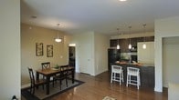 This single-floor residence offers a thoughtfully designed open-concept layout that blends functionality with modern style. The kitchen is a standout feature, outfitted with dark espresso cabinetry, black appliances, a tile backsplash, and a breakfast bar with counter-height seating — perfect for casual dining or entertaining. Pendant lighting hangs above both the kitchen island and the adjacent dining area, adding a polished, contemporary touch. The dining space comfortably accommodates a full table and chair set, anchored by a woven area rug atop warm hardwood flooring that extends throughout the main living areas. Neutral-toned walls provide a clean, versatile backdrop, while framed wall art adds a welcoming character. A central hallway connects the open living area to additional rooms beyond, hinting at a practical and well-organized floor plan. The overall ambiance is warm and inviting, with a cohesive design aesthetic that balances modern finishes with everyday comfort. Natural light filters through the space, enhancing the airy feel of the open layout and highlighting the rich tones of the hardwood floors.