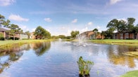 This single-story residence is situated within a well-maintained community that offers a notably tranquil outdoor setting. The most striking feature of the surrounding grounds is a large, calm pond positioned prominently within the community's landscape. Two decorative fountains rise from the water's surface, creating elegant cascading sprays that add both visual interest and a sense of liveliness to the serene environment. The pond's reflective surface mirrors the open sky above, enhancing the overall sense of space and openness.

The community grounds are generously landscaped with expansive stretches of well-kept grass and mature trees that provide natural shade and greenery throughout. Multi-story residential buildings with warm earth-toned exteriors are visible in the background, framing the water view and contributing to the cohesive character of the development. The setting suggests a peaceful, park-like atmosphere that residents can enjoy from common outdoor areas.

While interior images were not sufficiently lit to provide detailed descriptions of the indoor spaces, the residence is contained within a single floor, offering the convenience and accessibility of single-level living within this attractively landscaped community environment.