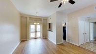 This single-floor residence offers an open and airy layout bathed in natural light. The main living area features warm hardwood flooring that extends throughout, contributing to a clean and cohesive aesthetic. Elevated ceilings enhance the sense of spaciousness, while a ceiling fan with a light fixture ensures year-round comfort. A decorative pendant chandelier adds a touch of elegance to the dining area.

A set of French doors serves as the focal point of the living space, opening to an outdoor balcony and filling the interior with abundant natural light. Transom windows above the doors further amplify the brightness of the room.

The kitchen is accessible via a convenient pass-through window with a breakfast bar, allowing for an open connection between the kitchen and living areas. Dark cabinetry in the kitchen provides a striking contrast to the neutral, warm-toned walls throughout the residence.

The overall color palette is soft and neutral, with beige walls creating a versatile backdrop suitable for a variety of interior styles. Floor vents are integrated discreetly into the layout. The space strikes a balance between functional design and inviting ambiance, making it a comfortable and well-appointed single-floor living environment.